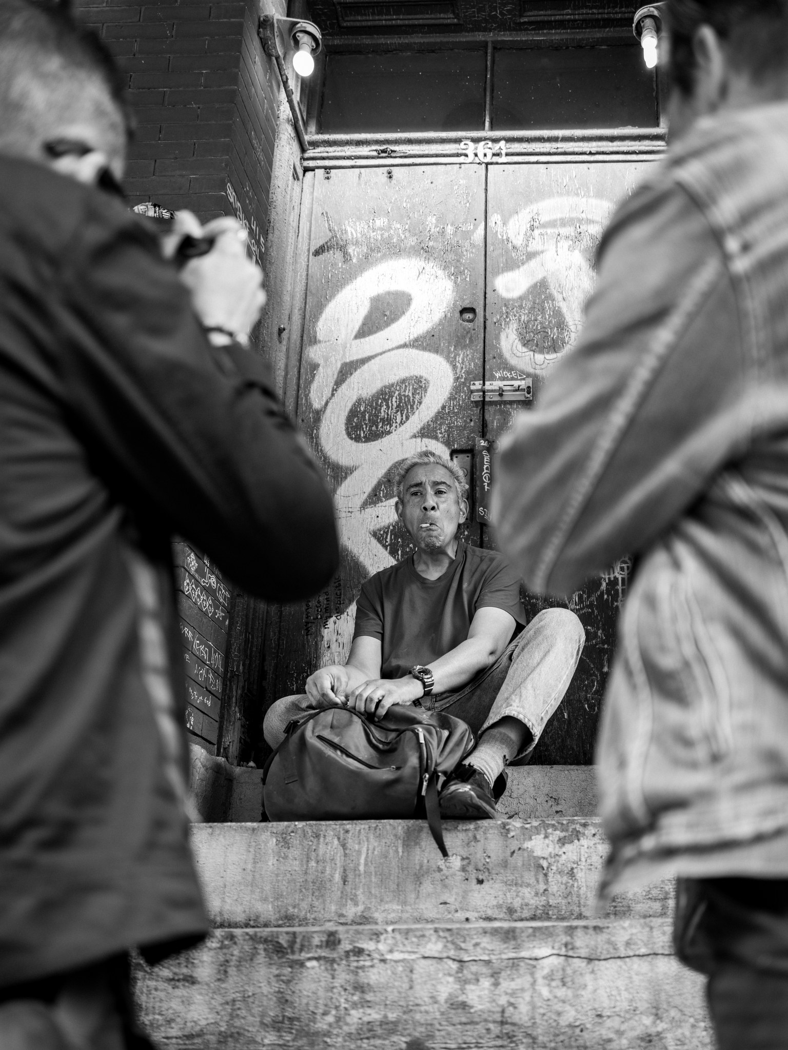 A man sitting at the top of concrete steps against a graffitied door numbered 361, a cigarette in his mouth and a bag in his lap, while two photographers seen from behind photograph him